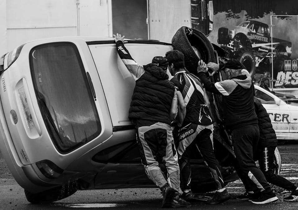 Quem é responsável em caso de acidente com motorista em treinamento? Black and white image of a team pushing an overturned car during a live stunt performance.