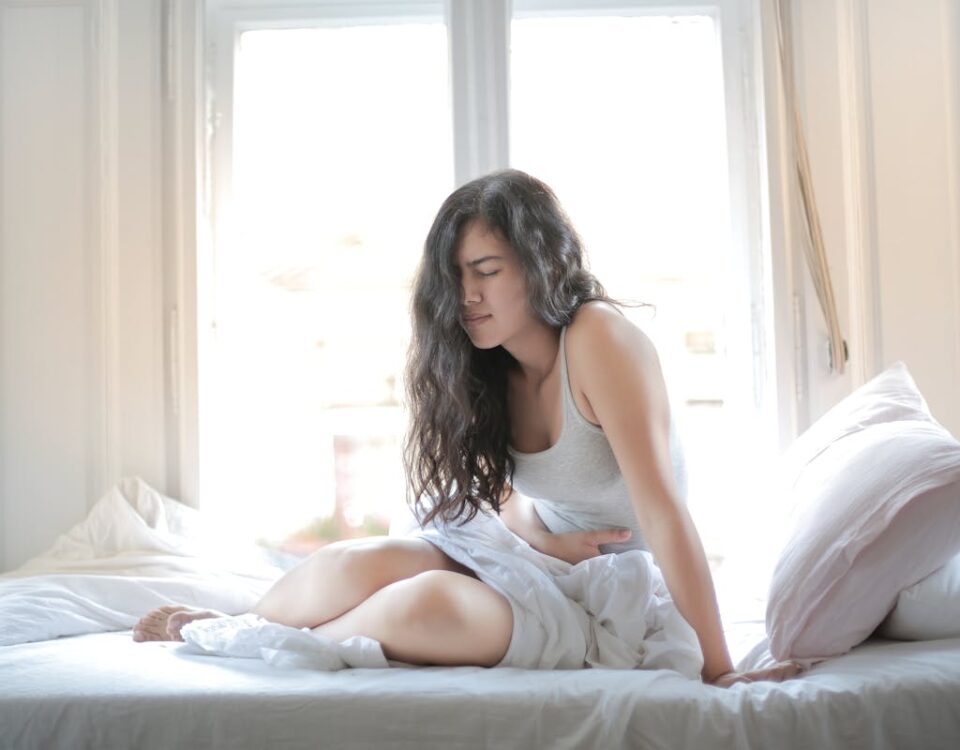 Cobertura obrigatória de DII pelos planos de saúde: o que saber Woman sitting on bed holding her stomach, showing signs of discomfort or pain indoors.
