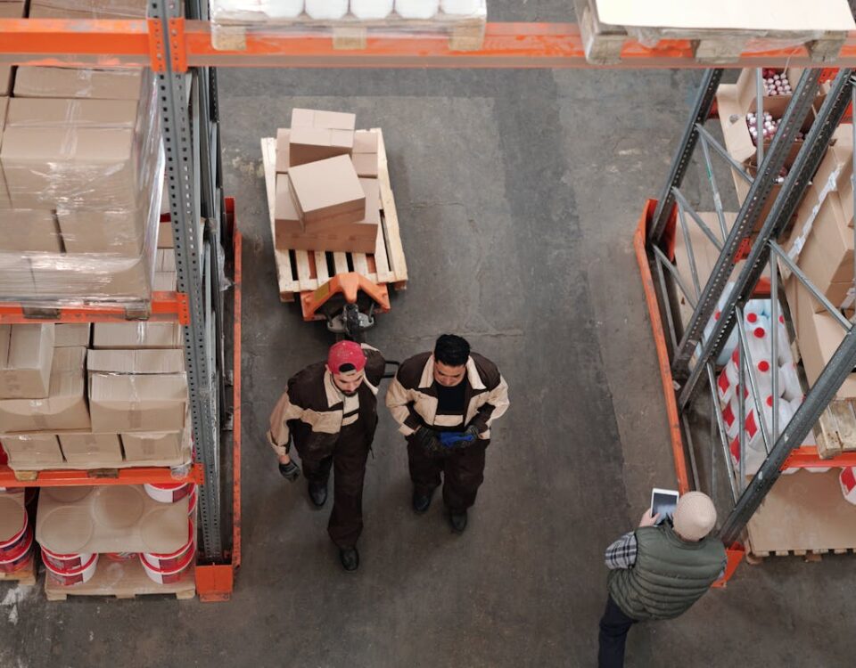 Quais regulamentações impactam os processos das grandes cadeias? High angle view of warehouse workers organizing and preparing shipments with boxes and pallets.