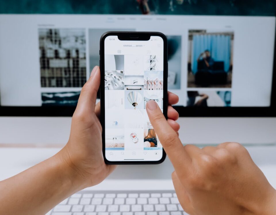 Hands holding a smartphone while browsing a social media app in front of a desktop computer.