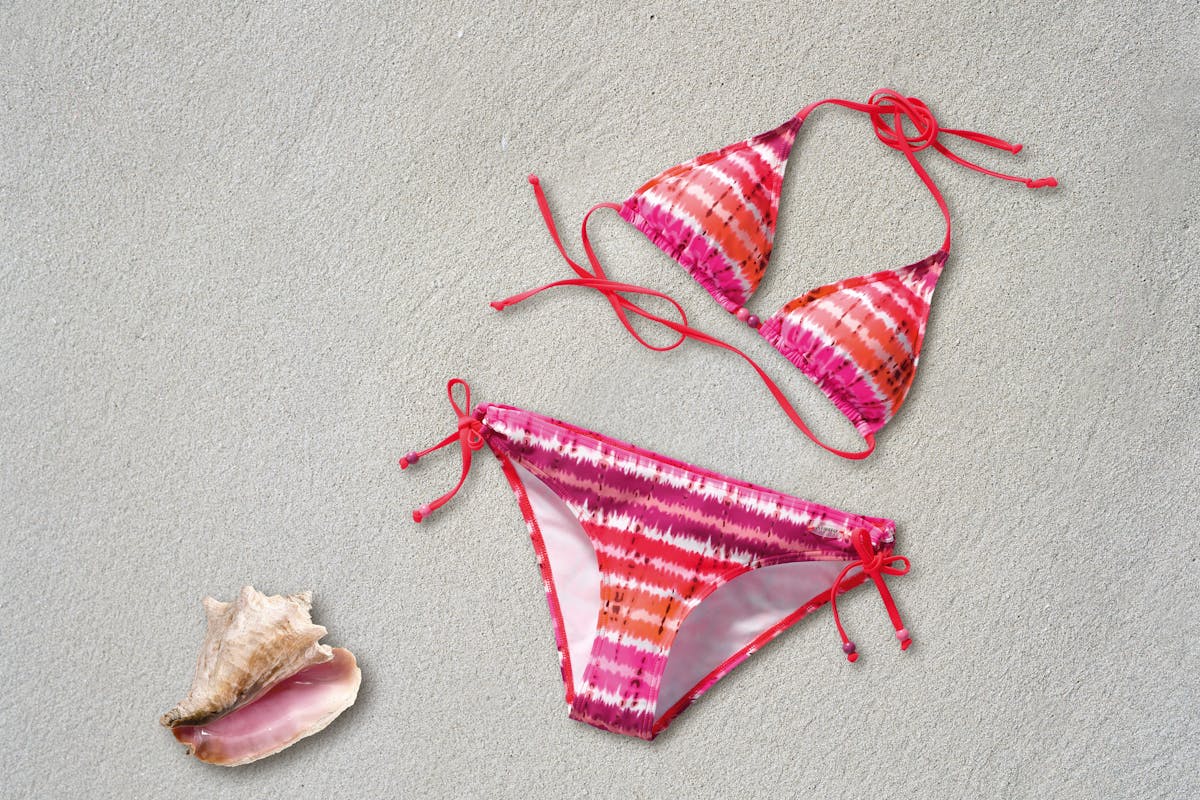 Colorful tie-dye bikini on sand next to a pink seashell, ideal for summer vibes.