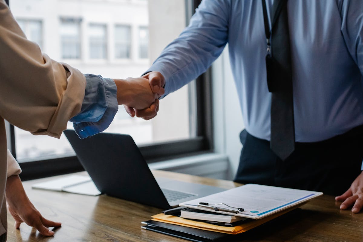 Entenda o processo de obtenção de licença ambiental Crop unrecognizable coworkers in formal wear standing at table with laptop and documents while greeting each other before meeting