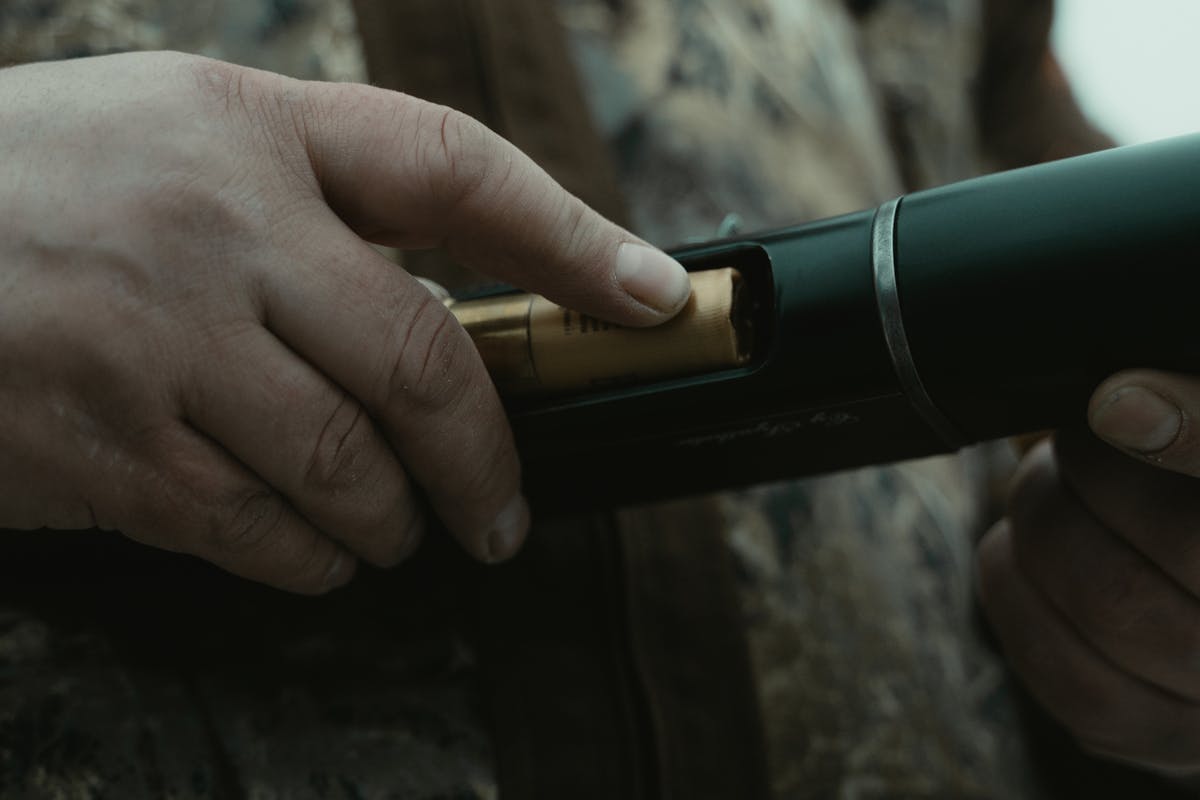 Close-up of a person inserting a bullet into a shotgun with camouflage attire in the background.