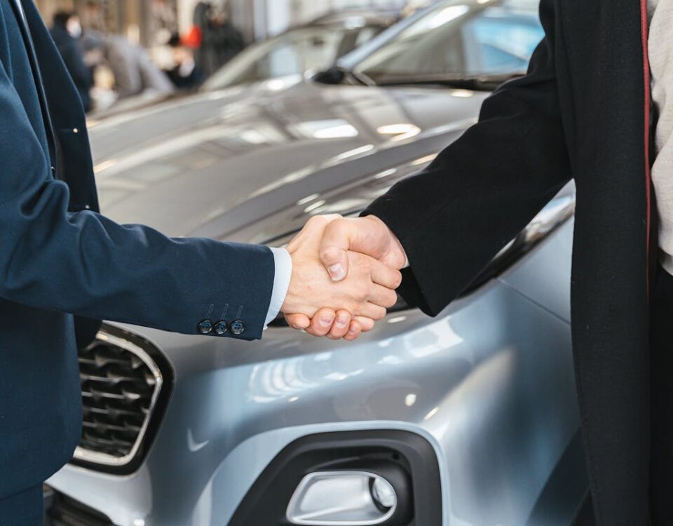 Direitos e deveres ao contratar um seguro de carro A close-up of two people shaking hands in front of a car in an indoor setting.