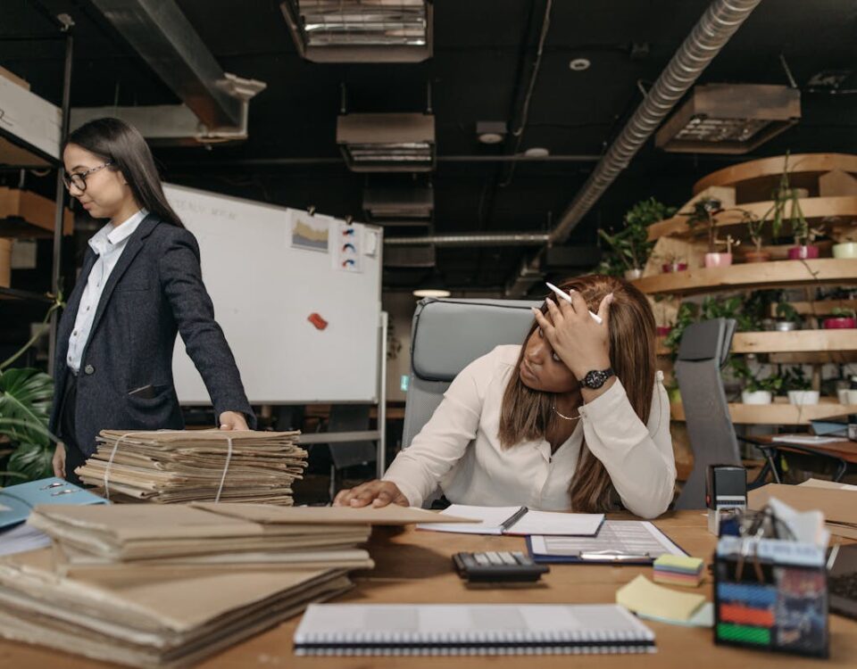 Quais são os direitos do auxiliar administrativo? Two women working with documents in a modern office setting, one sitting at the desk, the other standing.