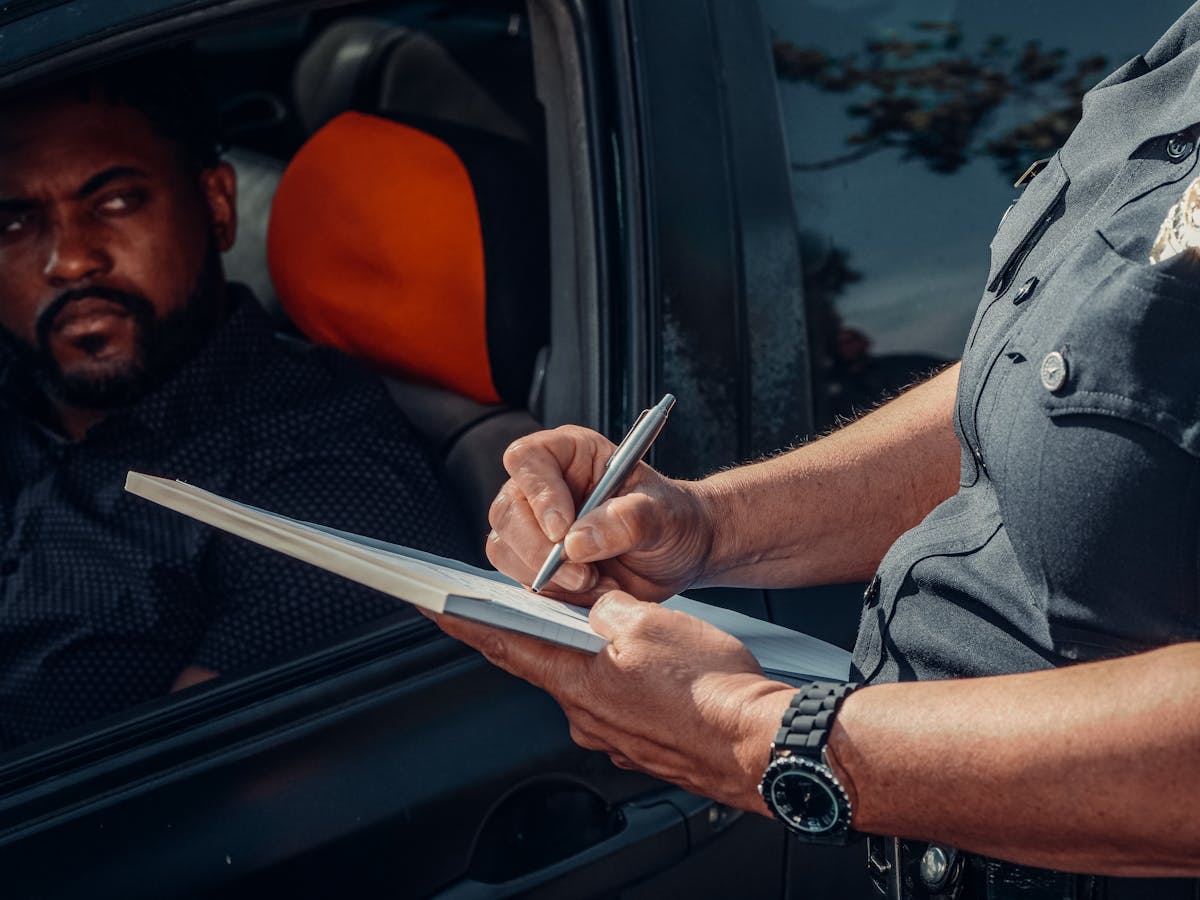 Quais infrações podem suspender sua CNH? A police officer writes a ticket as the driver looks on from inside the car.