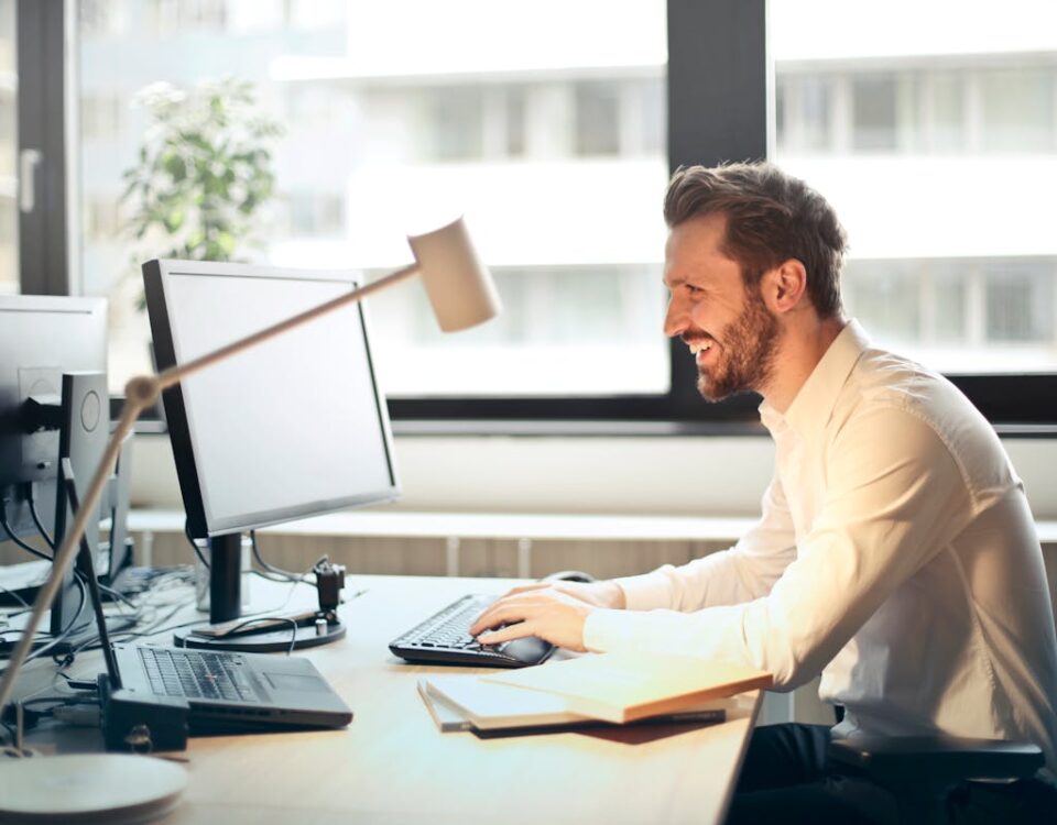 Automação comercial e compliance: O que sua empresa precisa saber? A man smiling while working at an office desk with a computer and natural daylight streaming in through large windows.