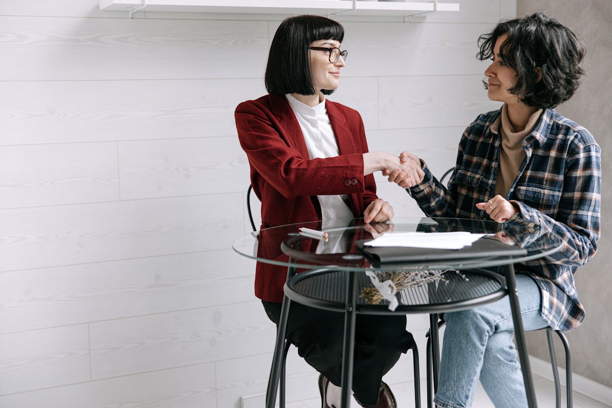 Two women shaking hands in a professional meeting, seated at a glass table indoors.