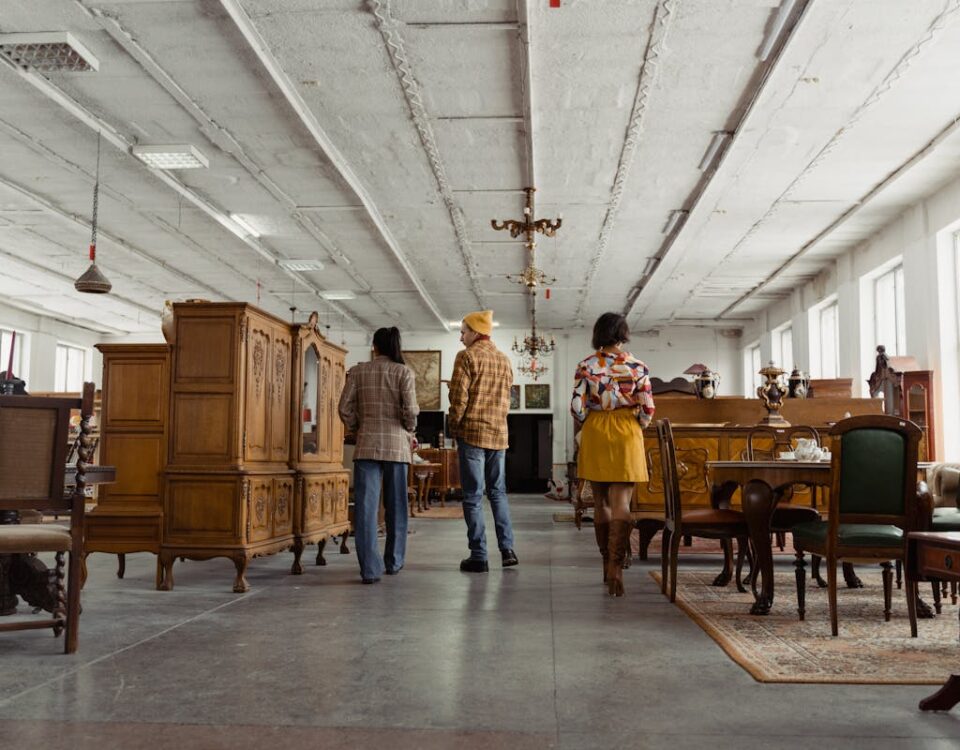 Direitos do consumidor na compra de móveis de luxo Three people browsing vintage wooden furniture in a spacious store interior with high ceilings.