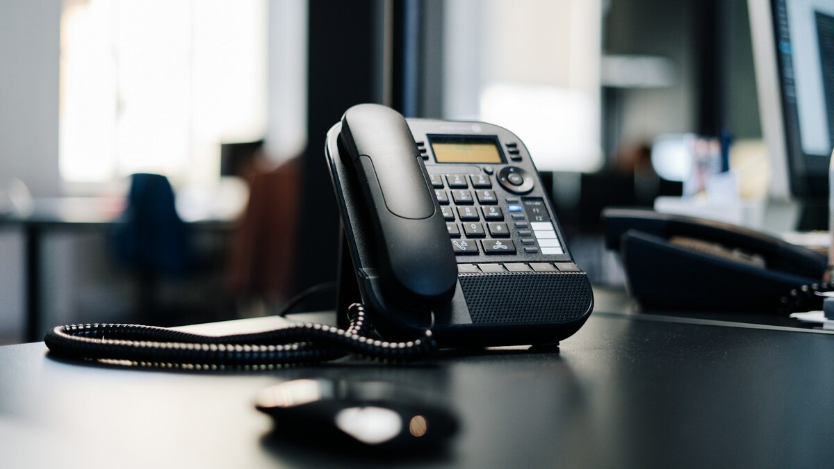 Telefone de mesa preto sobre bancada de escritório com computador ao fundo