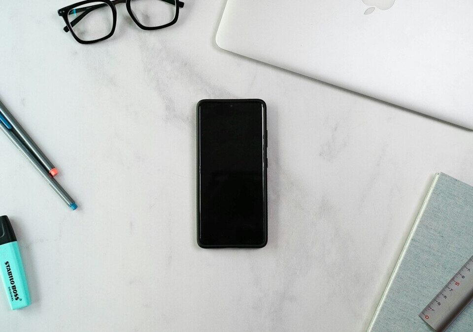 Celular sobre mesa de escritório branca ao lado de notebook, canetas e caderno