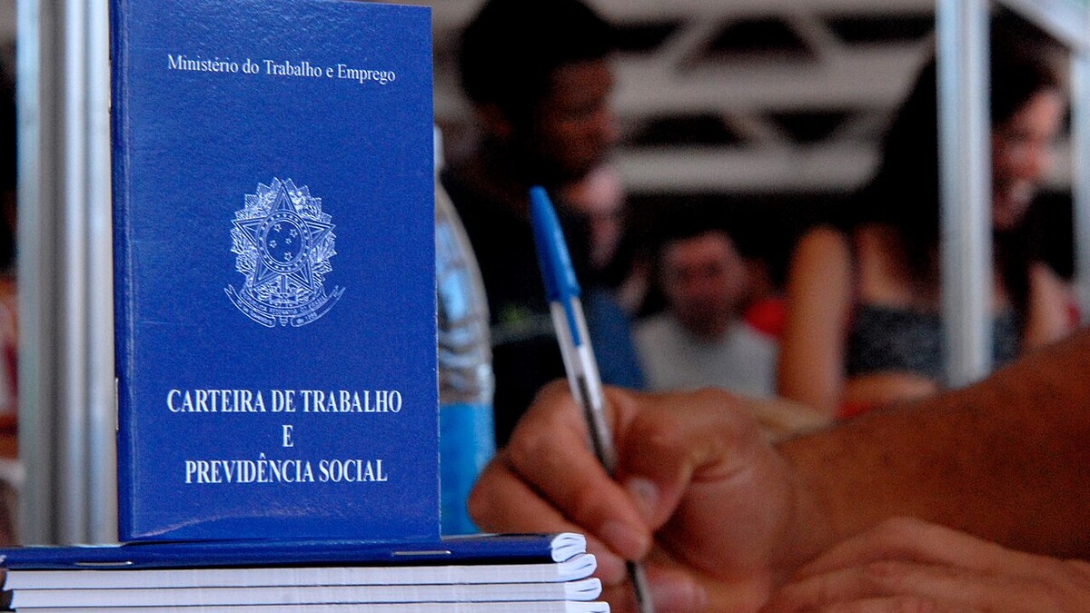 Carteira de Trabalho e Previdência Social sobre uma mesa, com pessoas preenchendo papéis ao fundo