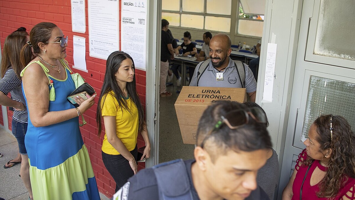 Eleitores em fila diante de uma seção eleitoral com urna sendo transportada